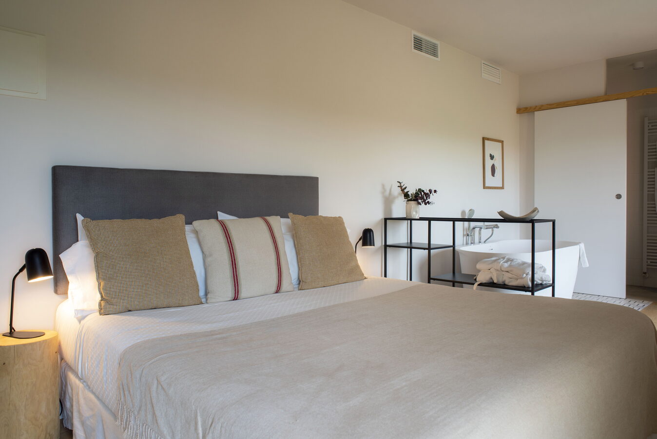 Llit de matrimoni amb capçal gris i banyera exempta al fons de la habitació