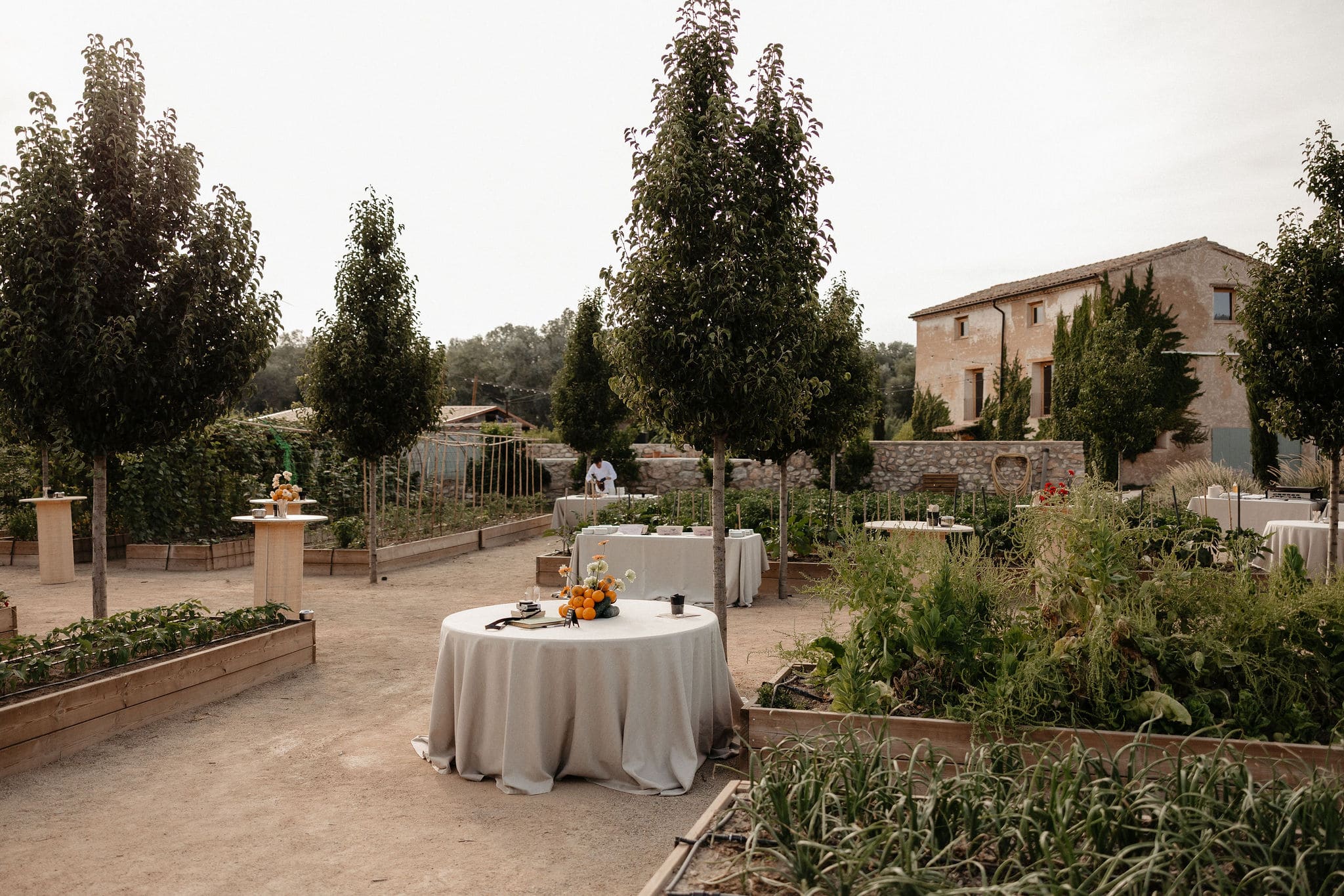 Taules amb tovalles clares a l'hort de la finca amb la masia al fons