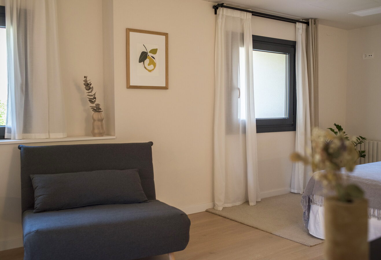 Habitació El Llimoner amb butaca grisa, làmina d'una llimona i finestra amb cortines