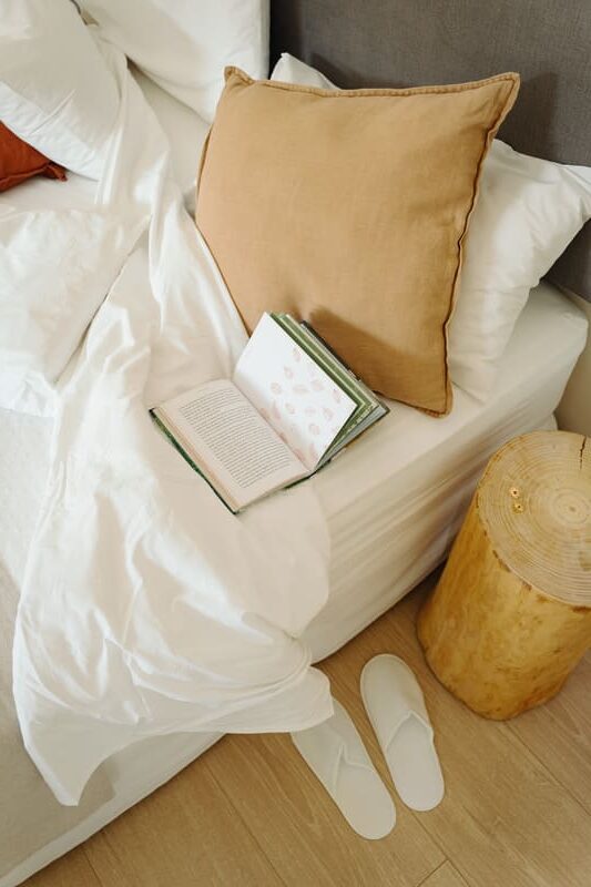 Llit desfet amb coixins de lli, llibre obert i sabatilles