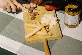 Mans tallant embotit i formatge sobre una taula de fusta amb pot d'olives
